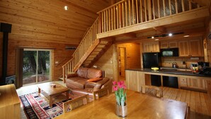 Interior de una cabaña de madera con sala de estar, comedor, cocina y una escalera que conduce a un nivel superior.