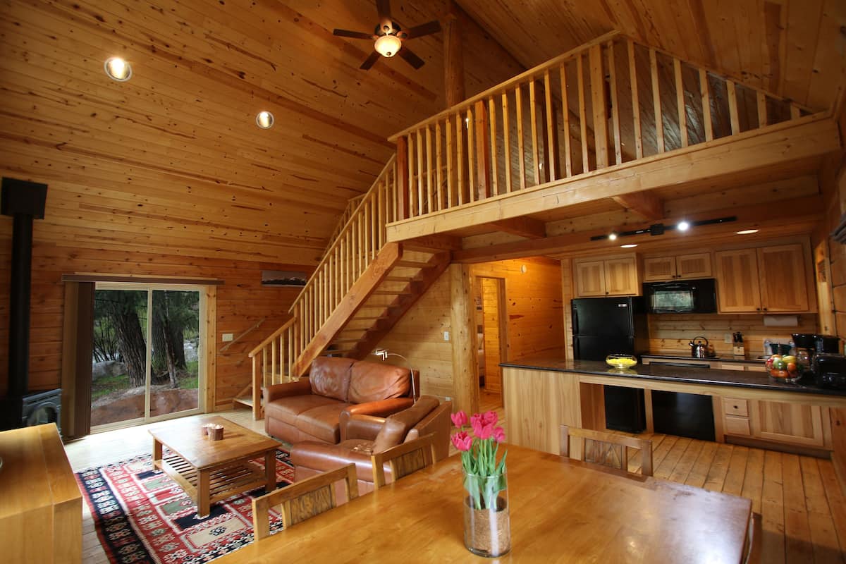Interior de una cabaña de madera con sala de estar, comedor, cocina y una escalera que conduce a un nivel superior.