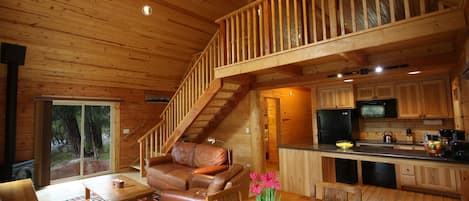 Interior de una cabaña de madera con sala de estar, comedor, cocina y una escalera que conduce a un nivel superior.