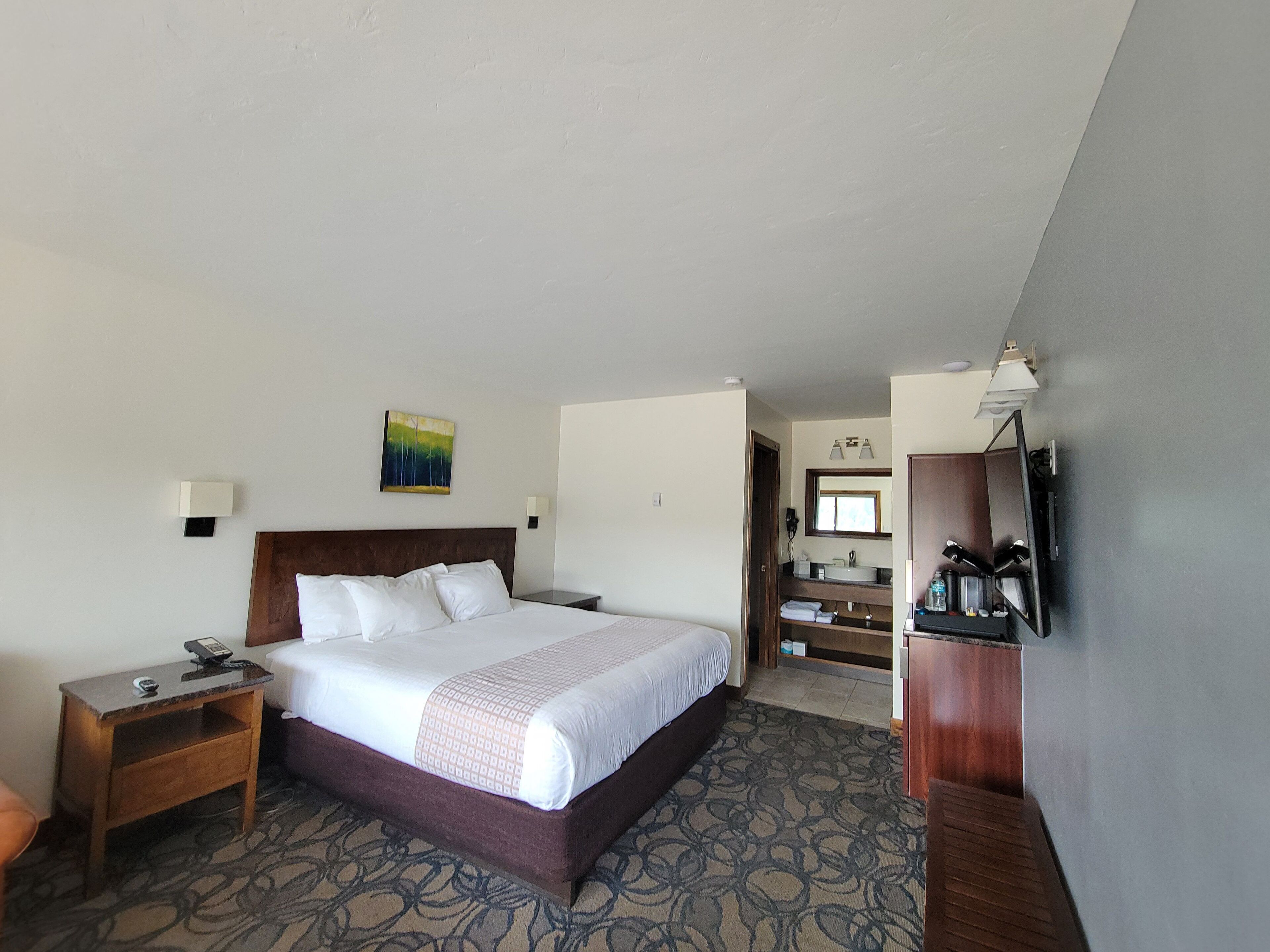 Ein Hotelzimmer mit einem großen Bett, einem Nachttisch, einem Schreibtisch, einem Fernseher und einem durch eine offene Tür einsehbarer Badezimmer.
