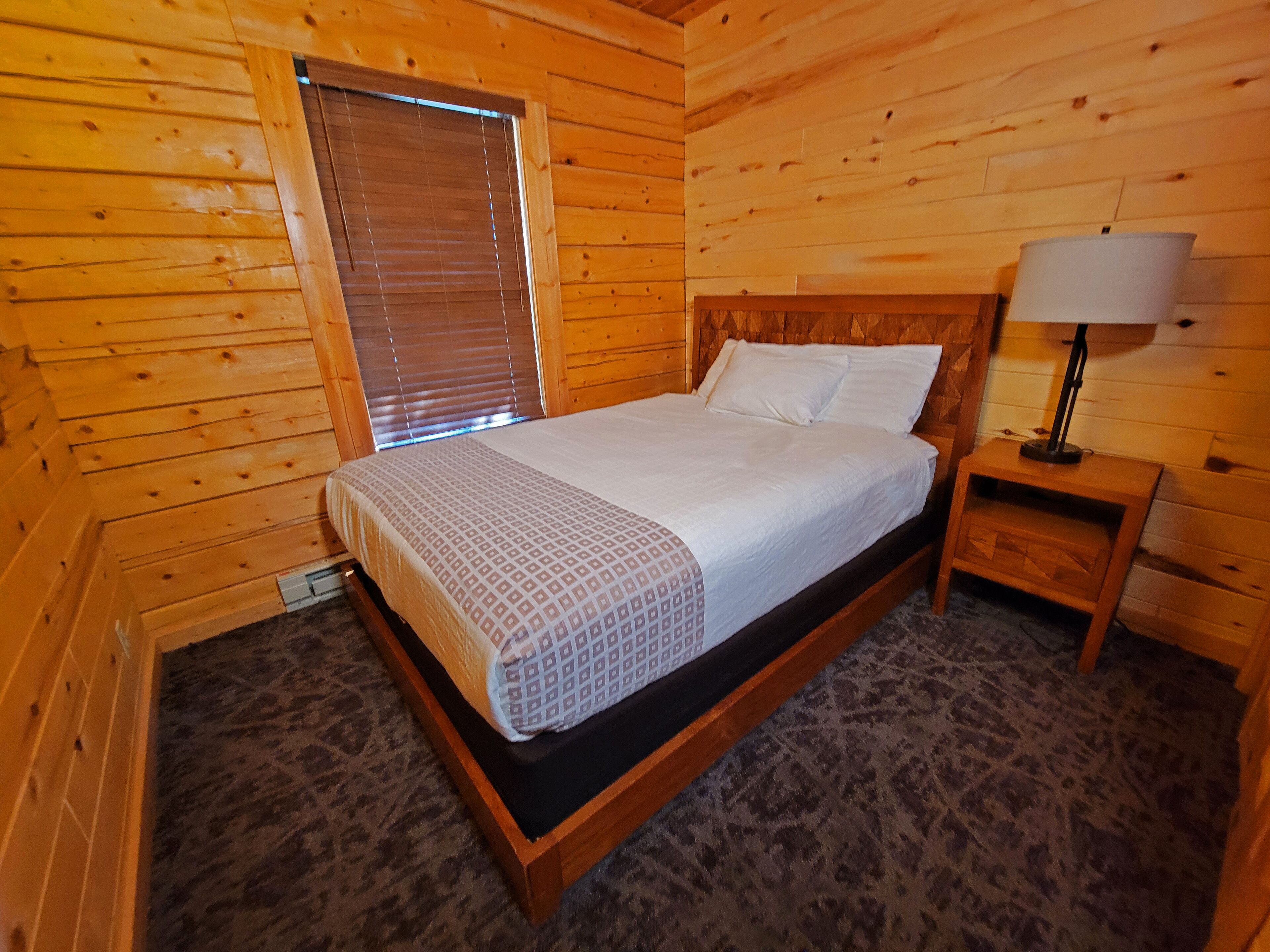 A wooden cabin bedroom with a bed, a nightstand, a lamp, and a window with blinds.