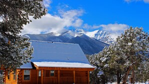 Una cabaña de madera con techo nevado, rodeada de árboles cubiertos de nieve y montañas al fondo.