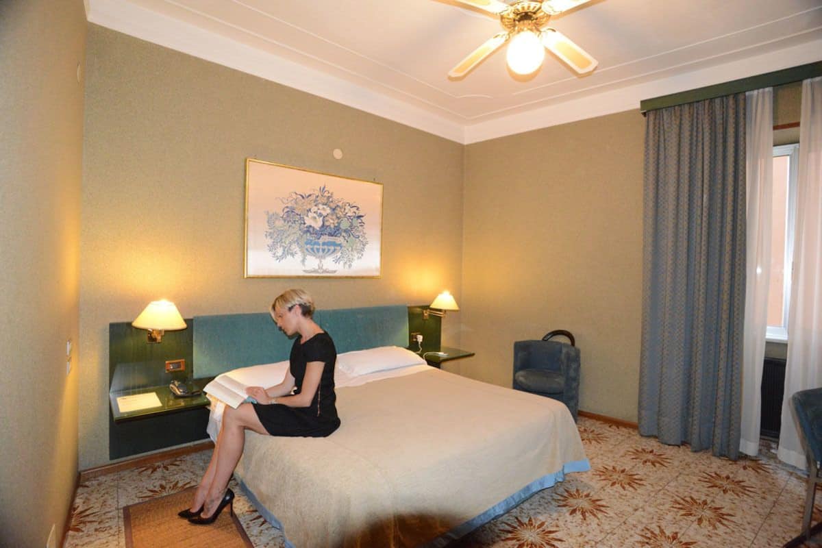 Una habitación de hotel con cama, dos lámparas de noche, ventilador de techo, un cuadro en la pared y una ventana con cortinas.