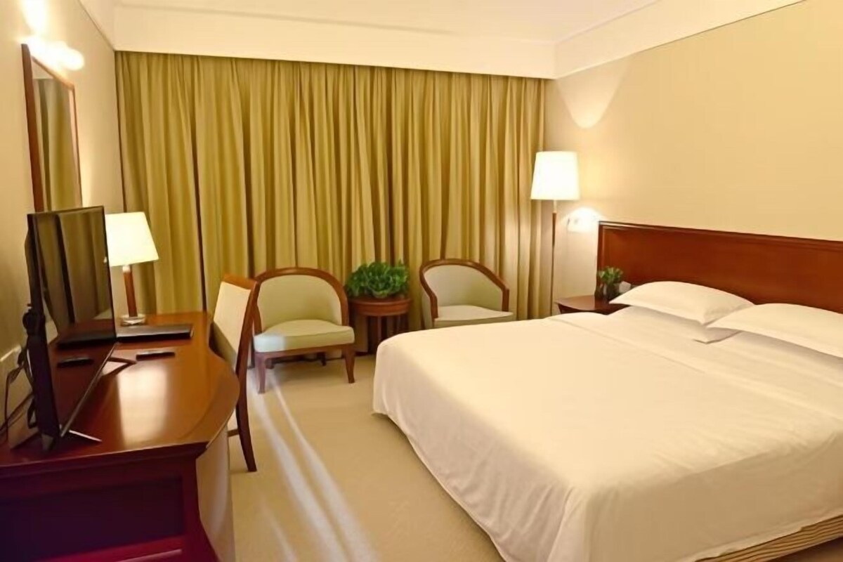 A hotel room with a bed, two chairs, a desk with a lamp, a mirror, and a plant.