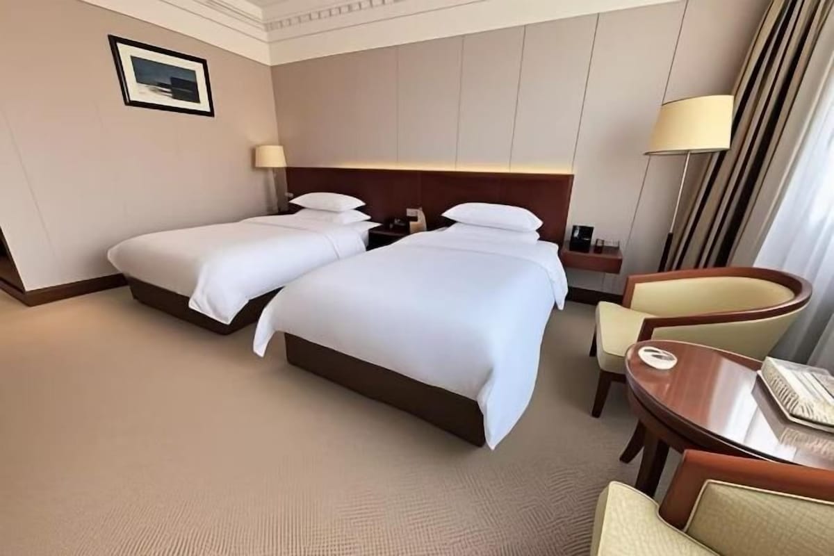 A hotel room with two beds, a desk, a chair, and a lamp.