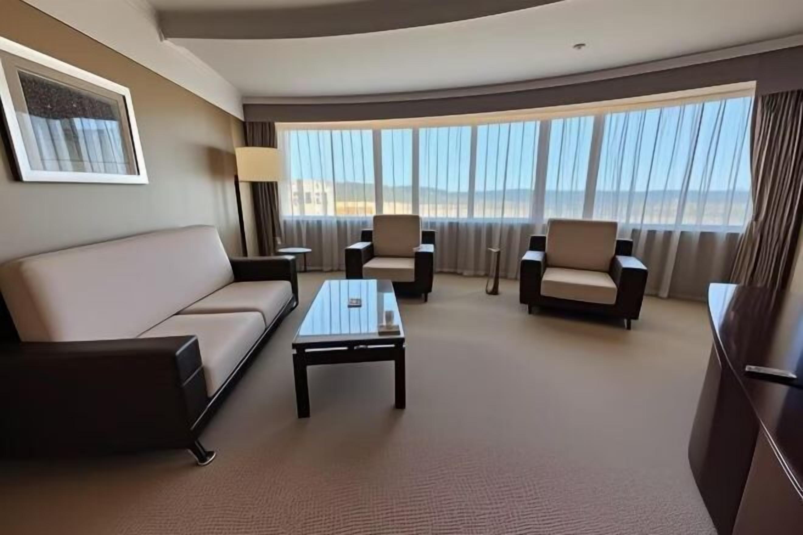 A hotel room with a sofa, two armchairs, a coffee table, a lamp, and a large window with a view.
