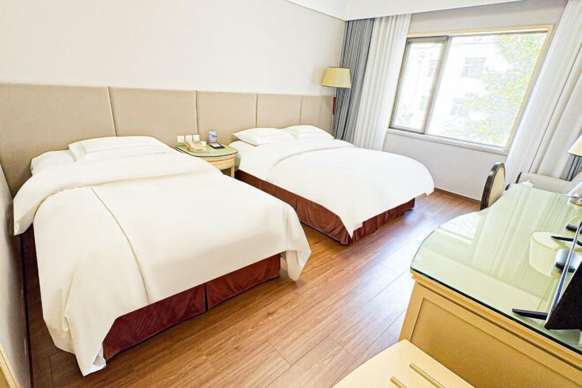 Une chambre d’hôtel avec deux lits, une table de chevet avec une lampe, un bureau avec un plateau en verre et une fenêtre avec des rideaux.