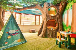 Ein Spielzimmer mit einem spielhaus im Baumdesign, einer Hängeschaukel und einem kleinen Tisch mit Spielzeug.
