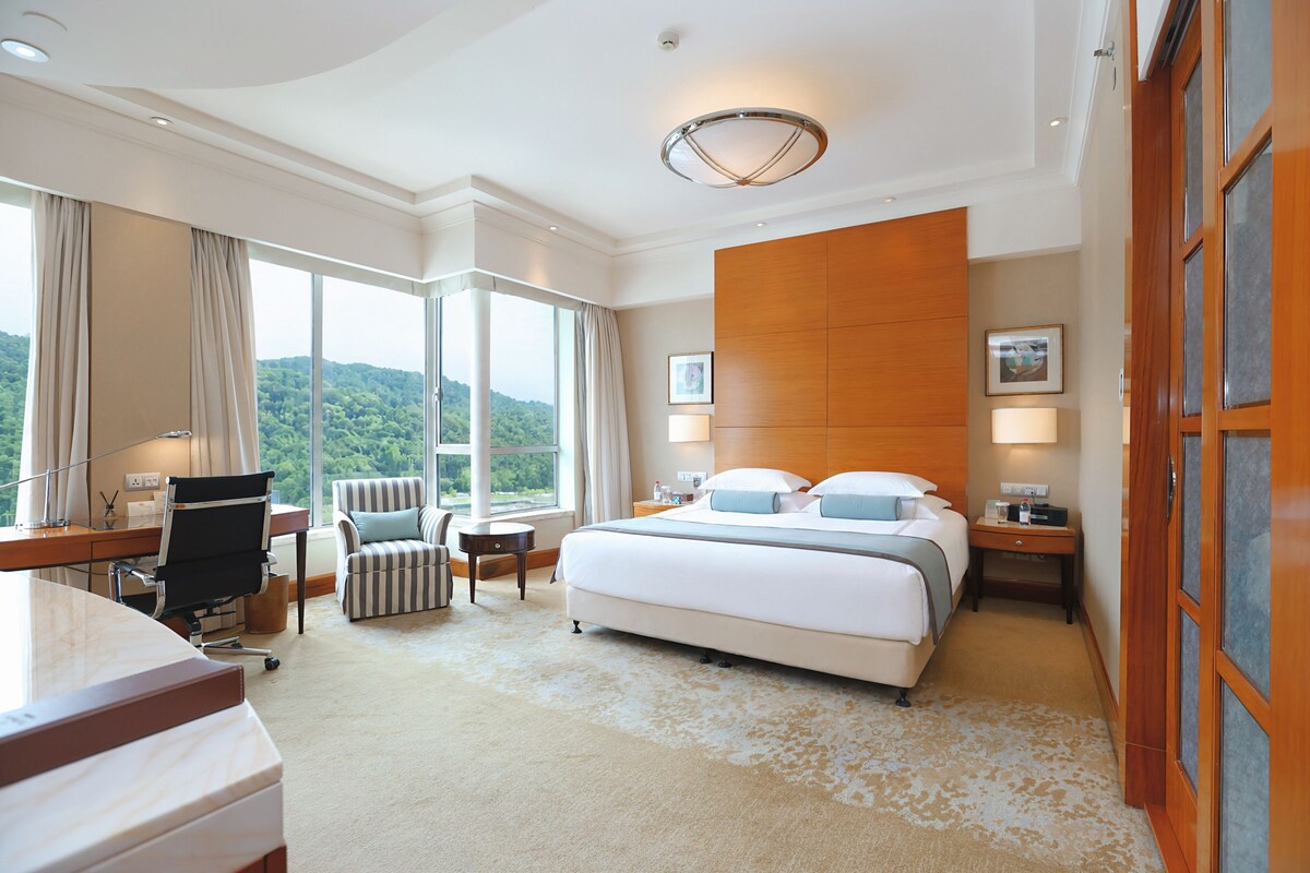A hotel room with a large bed, a desk, a chair, a view of the outdoors, and a wooden panel wall.