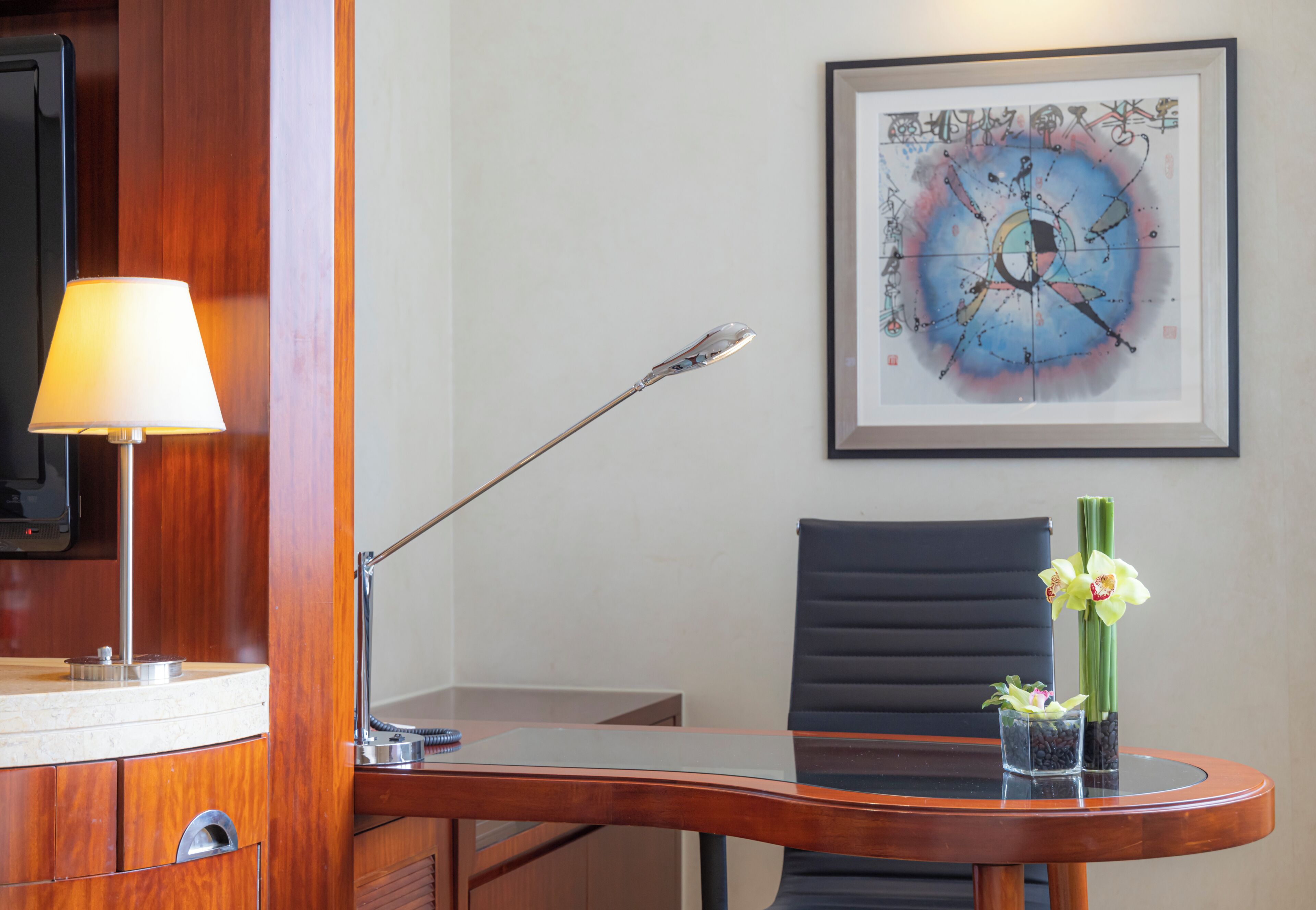 A hotel room with a desk, a chair, a lamp, a framed abstract painting, and a vase with flowers.
