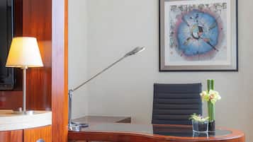 A hotel room with a desk, a chair, a lamp, a framed abstract painting, and a vase with flowers.