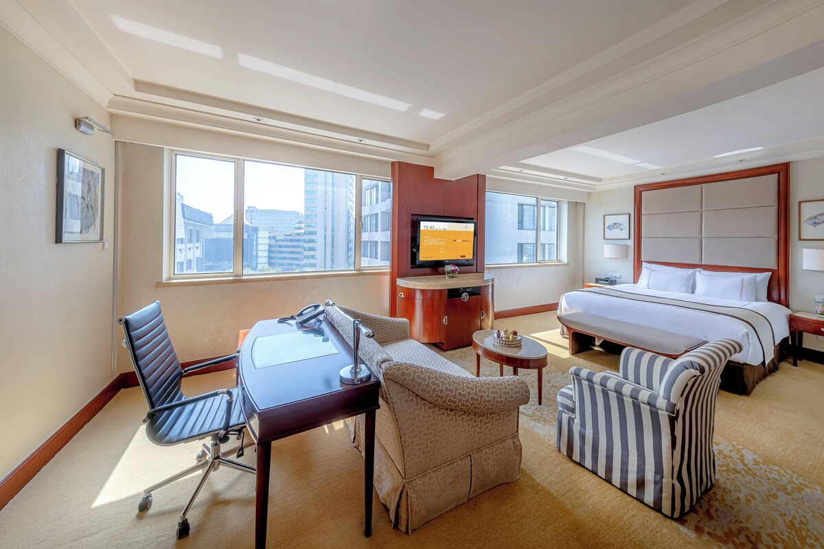 A hotel room with a bed, a desk with a chair, a sofa, a TV, and a window with a city view.