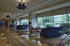 Eine geräumige Lobby mit poliertem Boden, einer Sitzecke mit Sofas und Sesseln, großen Fenstern mit Blick ins Grüne und einem Kronleuchter.