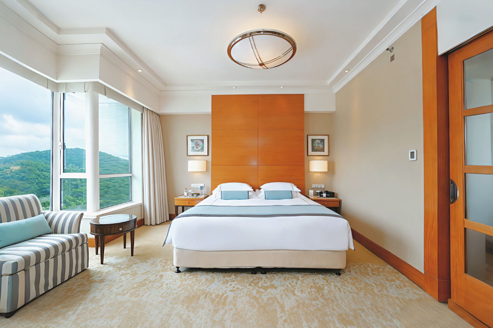 A hotel room with a large bed, a sitting area with a sofa, a view of the outdoors through a window, and a wooden door.