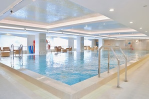 An indoor swimming pool with clear water, surrounded by lounge chairs and a sunbed.