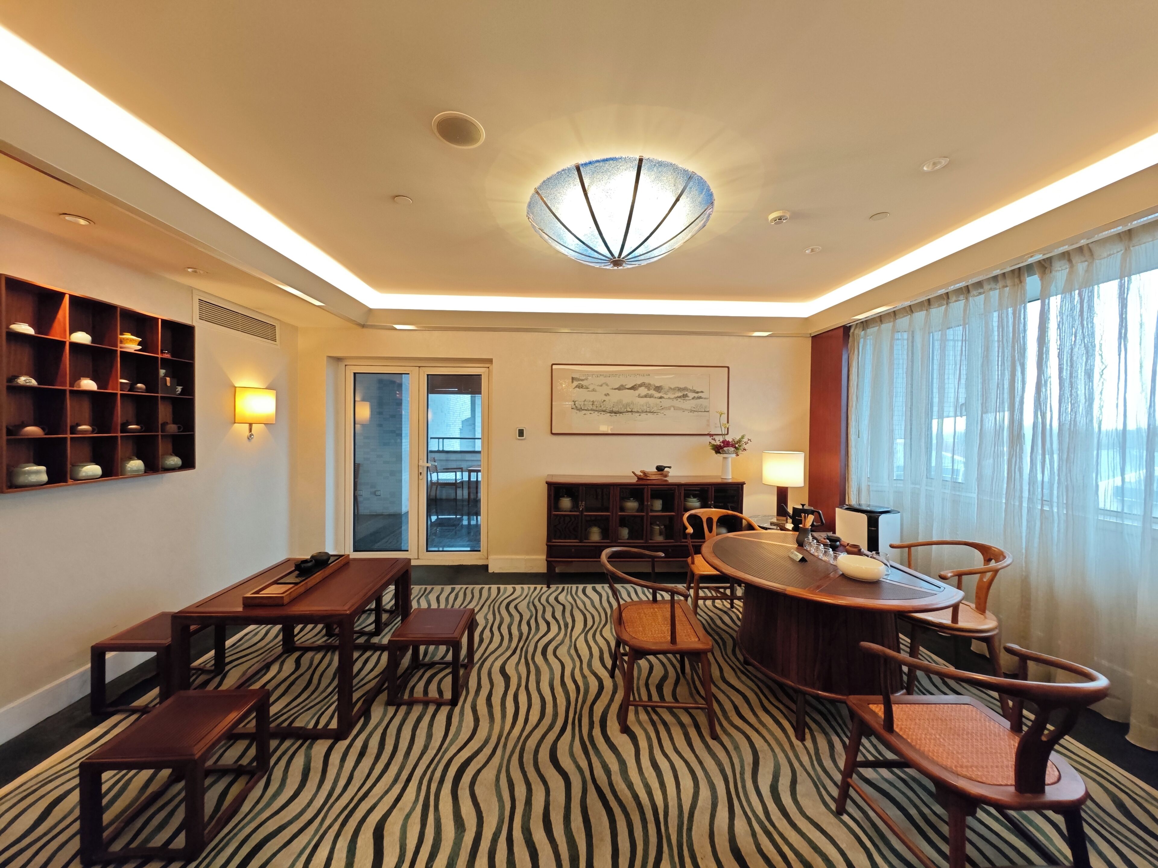 A hotel room with a dining area, a patterned carpet, a ceiling light fixture, and a view through the window.