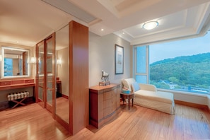A hotel room with a large window, wooden flooring, a wardrobe, a bedside table with a lamp, and a view of a forested area.