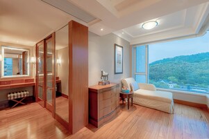 Habitación de hotel con un ventanal grande, suelo de madera, un armario, una mesita de noche con lámpara y vistas a zona boscosa.