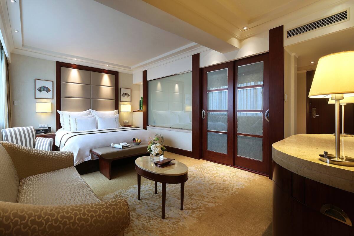 A hotel room with a bed, a sofa, a small table with a vase, a lamp, and a large window.