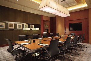 Un salón de conferencias con una mesa larga, múltiples sillas, un televisor, cuadros en la pared y una planta en maceta.