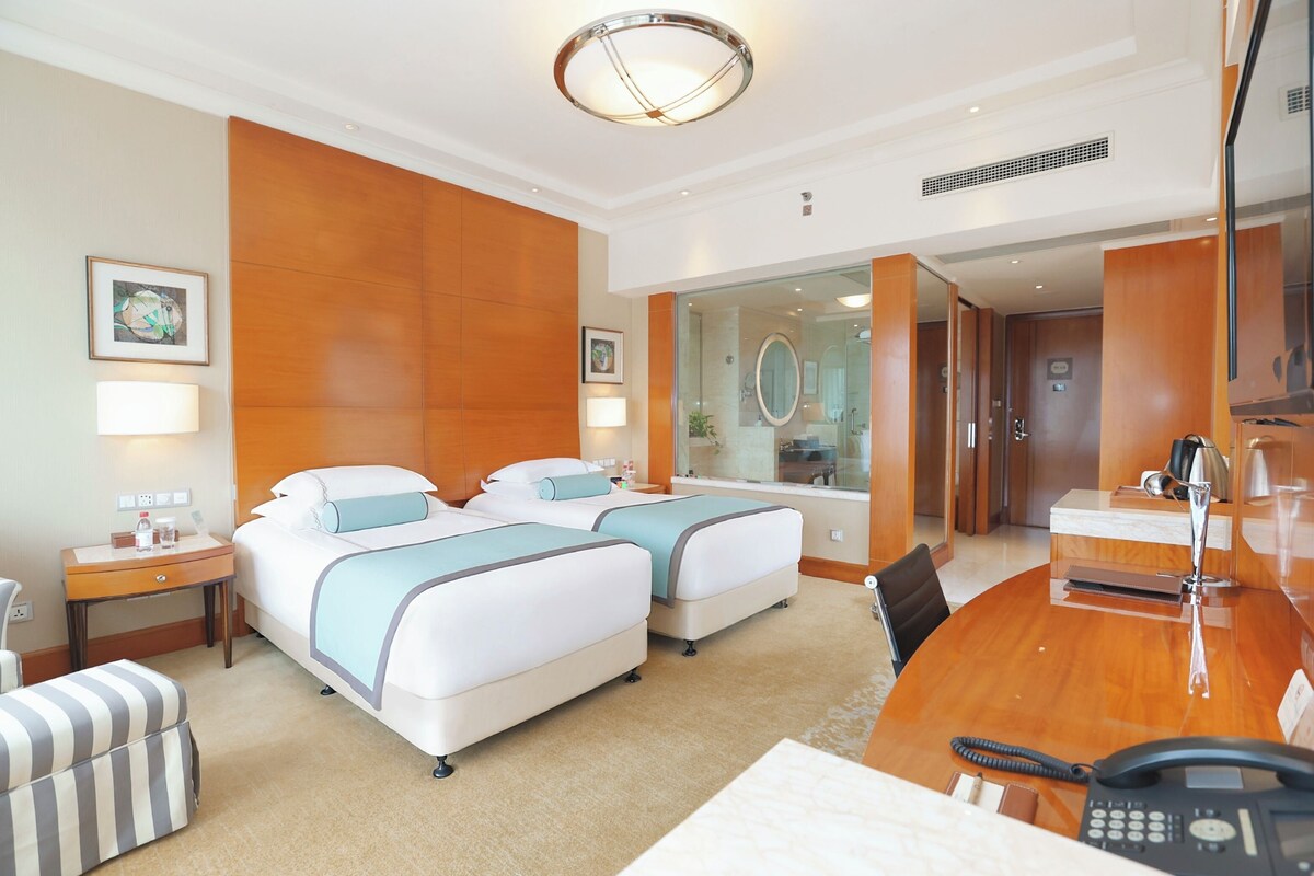A hotel room with two beds, a desk, a chair, a telephone, and a bathroom visible through a glass partition.
