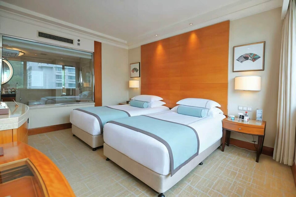 A hotel room with two beds, a desk, a mirror, and a window with a view of buildings.