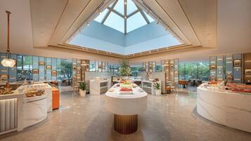 A spacious hotel lobby with a high ceiling, a central island with food displays, and a view of the outdoors through large windows.