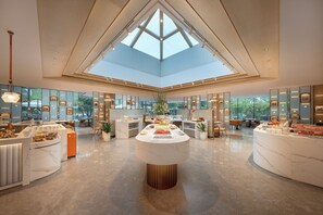 Een ruime hotellobby met een hoge plafond, een centraal eiland met etalages voor eten, en uitzicht op het buitenlandschap door grote ramen.