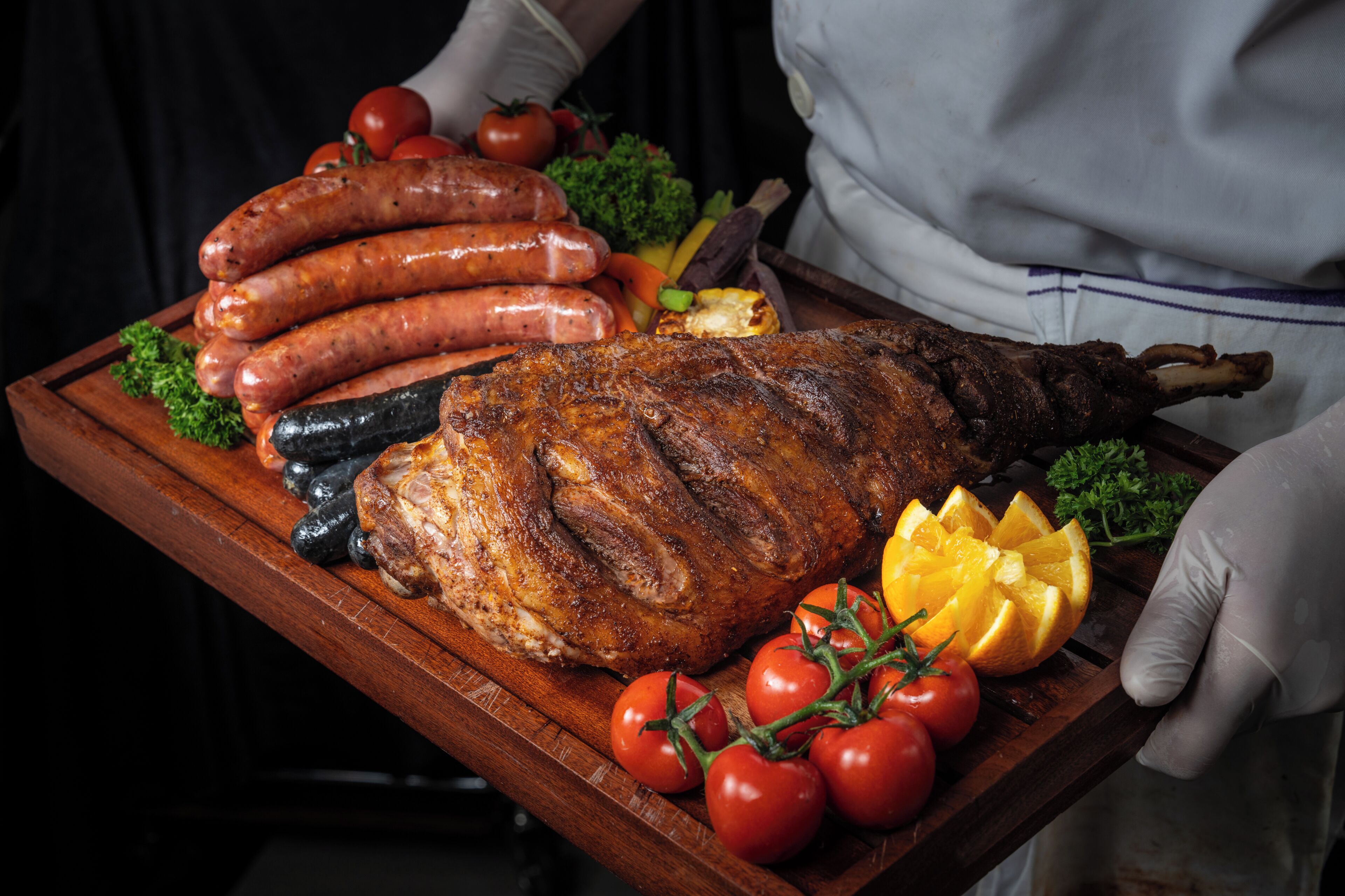 A roasted leg of lamb with sausages and tomatoes on a wooden board.