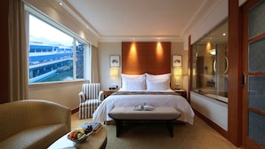 A hotel room with a large bed, a bedside table, a chair, a window with a view of buildings, and a bathroom with a glass door. - The Dragon Hotel (Hangzhou)