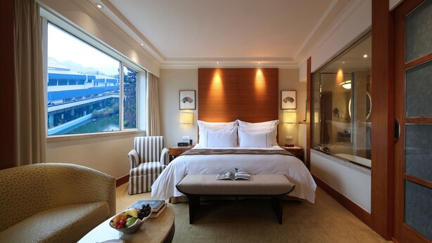 A hotel room with a large bed, a bedside table, a chair, a window with a view of buildings, and a bathroom with a glass door. - The Dragon Hotel (Hangzhou)