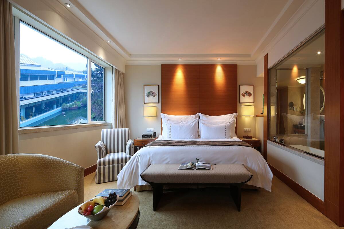 A hotel room with a large bed, a bedside table, a chair, a window with a view of buildings, and a bathroom with a glass door.