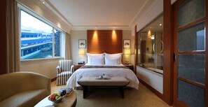 A hotel room with a large bed, a bedside table, a chair, a window with a view of buildings, and a bathroom with a glass door. - The Dragon Hotel (Hangzhou)
