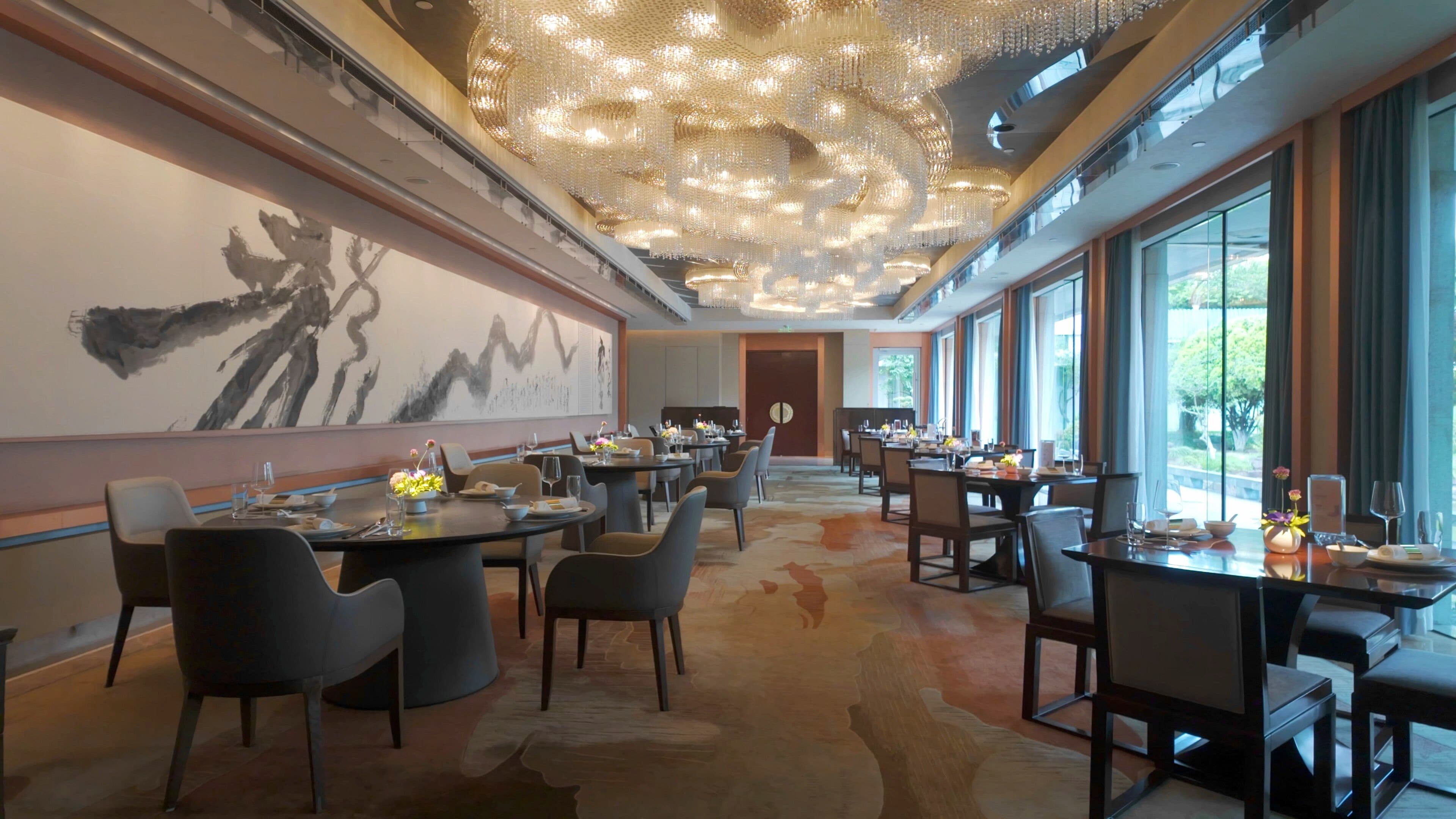 A spacious dining area with multiple tables and chairs, a large abstract painting on the wall, and a ceiling with intricate light fixtures.