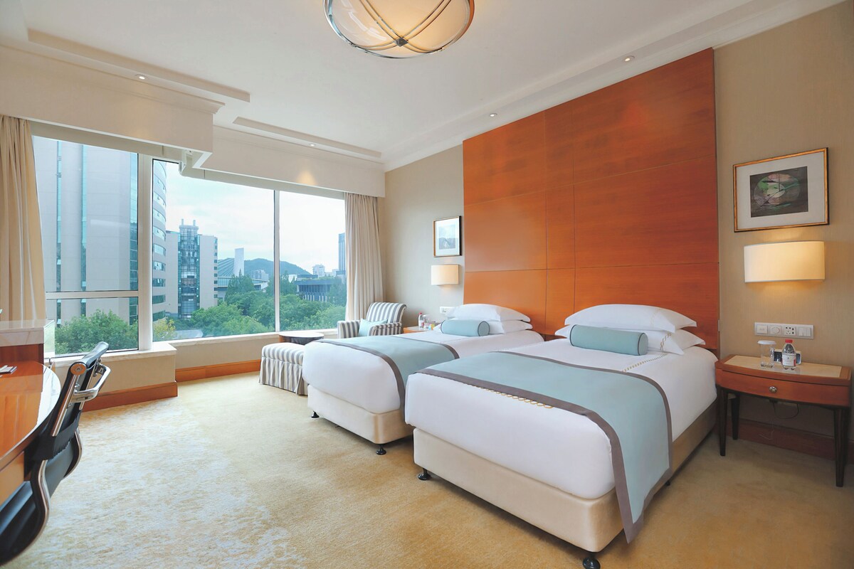 A hotel room with two beds, a desk, a chair, a large window with a city view, and a wall-mounted lamp.