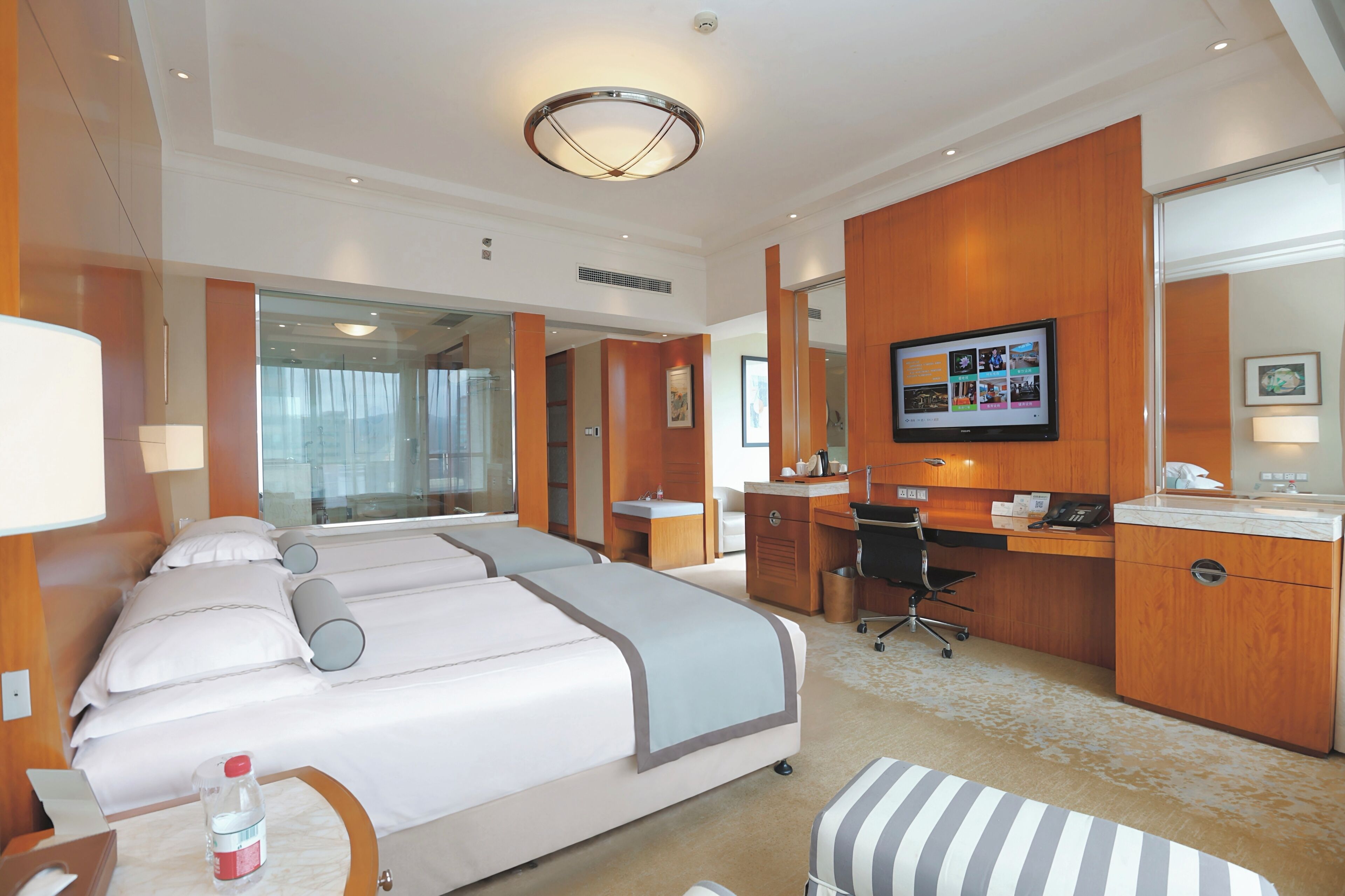 A hotel room with a large bed, a desk, a television, and a bathroom visible through a glass partition.