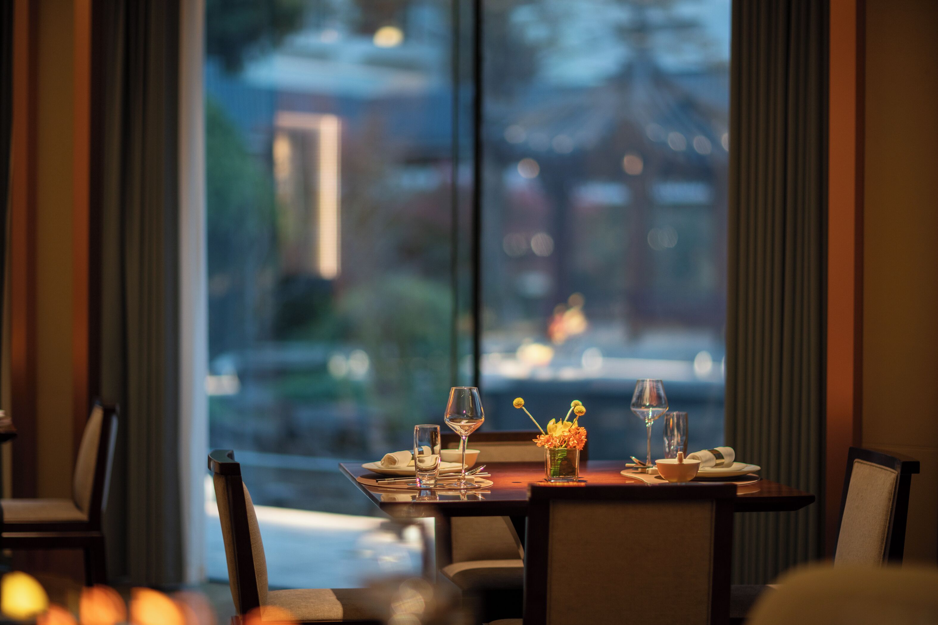 Una mesa de comedor dispuesta con platos, vasos y cubiertos, con vista a un paisaje urbano a través de amplios ventanales.