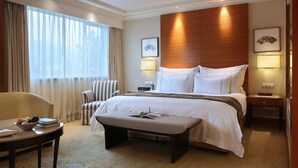A hotel room with a large bed, a bedside table, a chair, a small table with a bowl of fruit, and a window with curtains. - The Dragon Hotel (Hangzhou)
