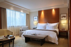 A hotel room with a large bed, a bedside table, a chair, a small table with a bowl of fruit, and a window with curtains. - The Dragon Hotel (Hangzhou)