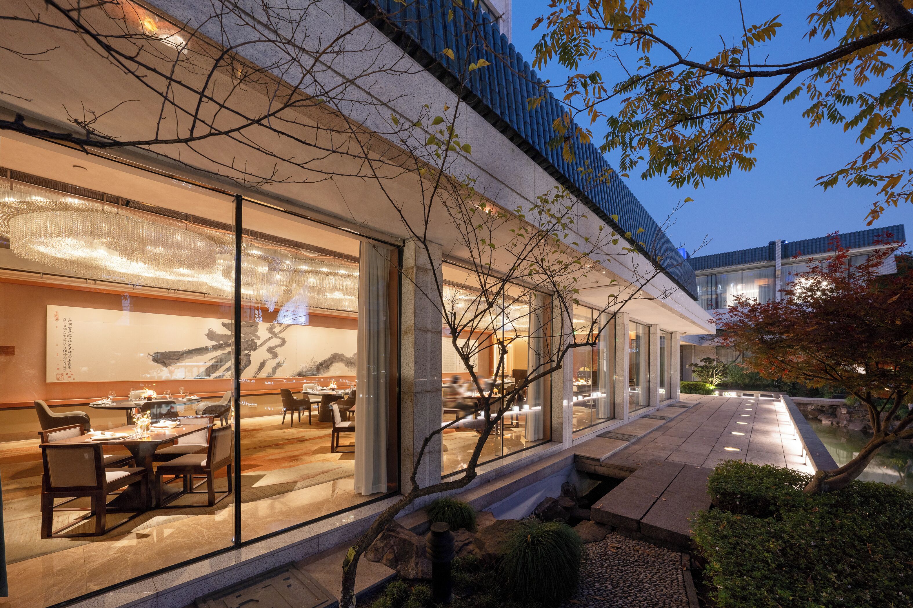 A modern building with large glass windows, a dining area with tables and chairs, and a water feature in the foreground.