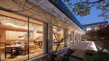 A modern building with large glass windows, a dining area with tables and chairs, and a water feature in the foreground.