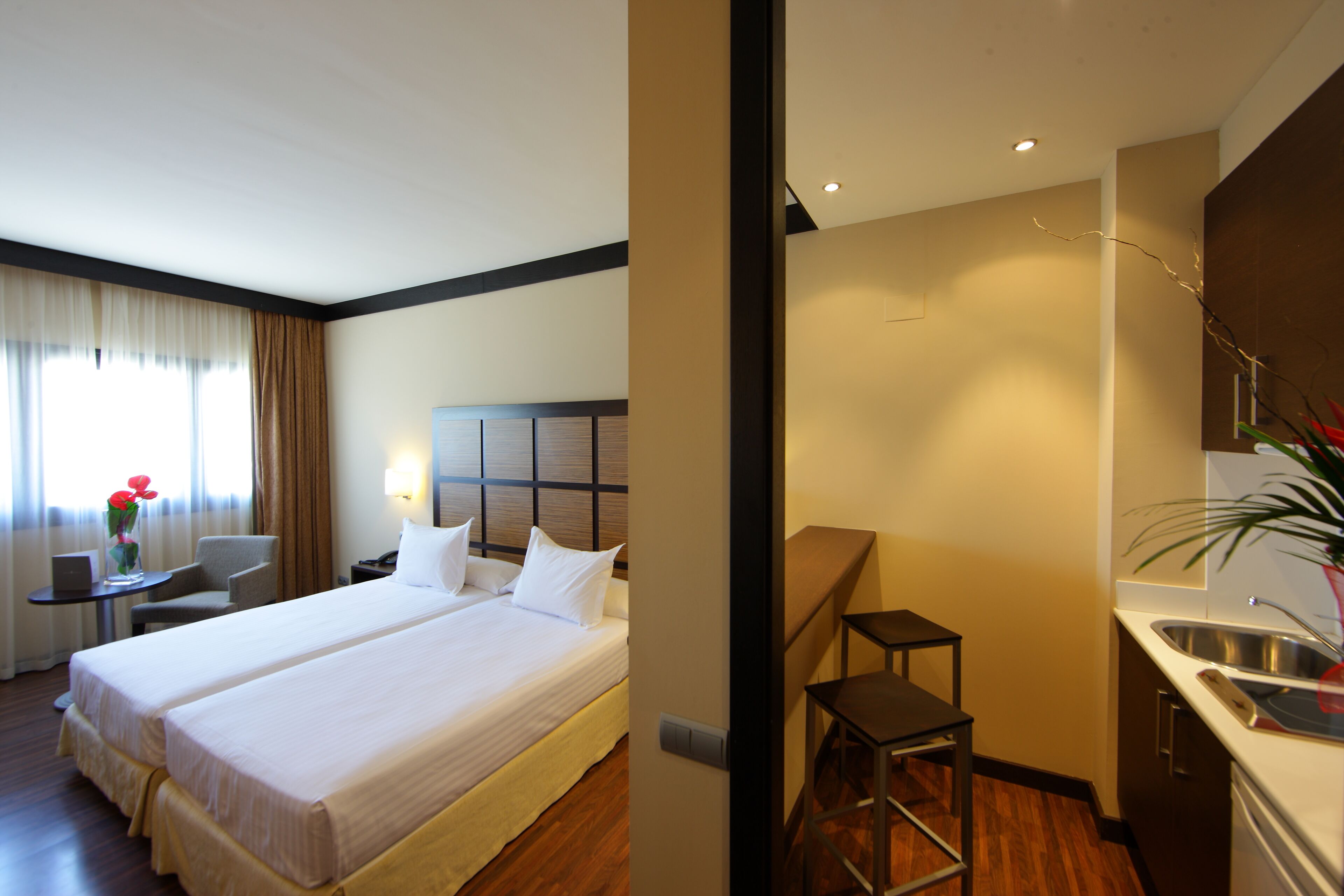 Une chambre d’hôtel avec deux lits, un bureau avec une chaise, une petite table avec un vase de fleurs, une table de chevet et une kitchenette équipée d’un évier et d’un plan de travail.