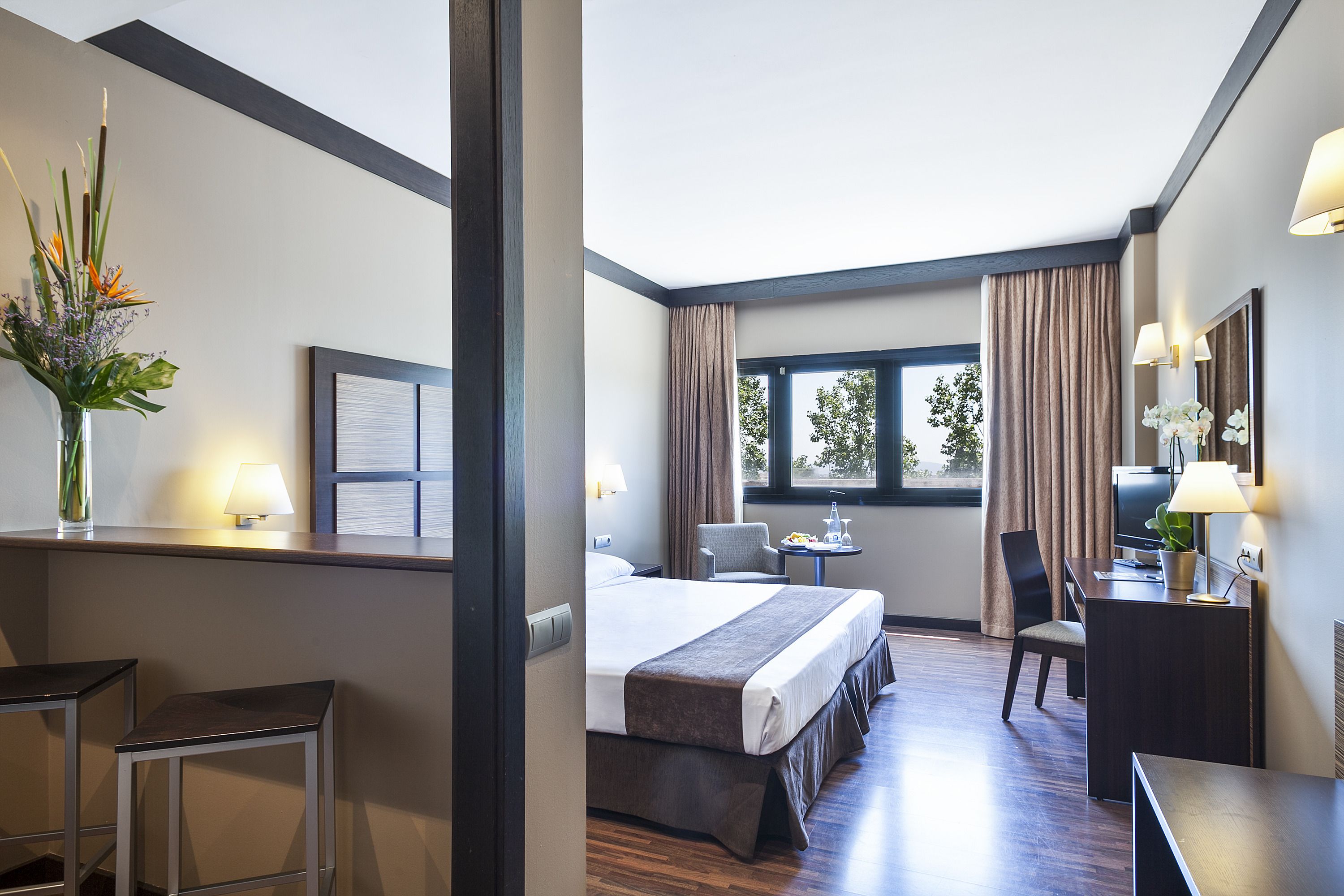A hotel room with a bed, a desk, two chairs, a small table, and a window with curtains.