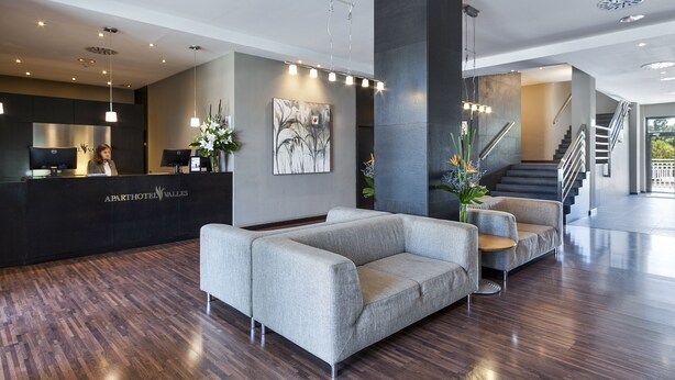 A modern hotel lobby with a reception desk, grey sofas, and a staircase.