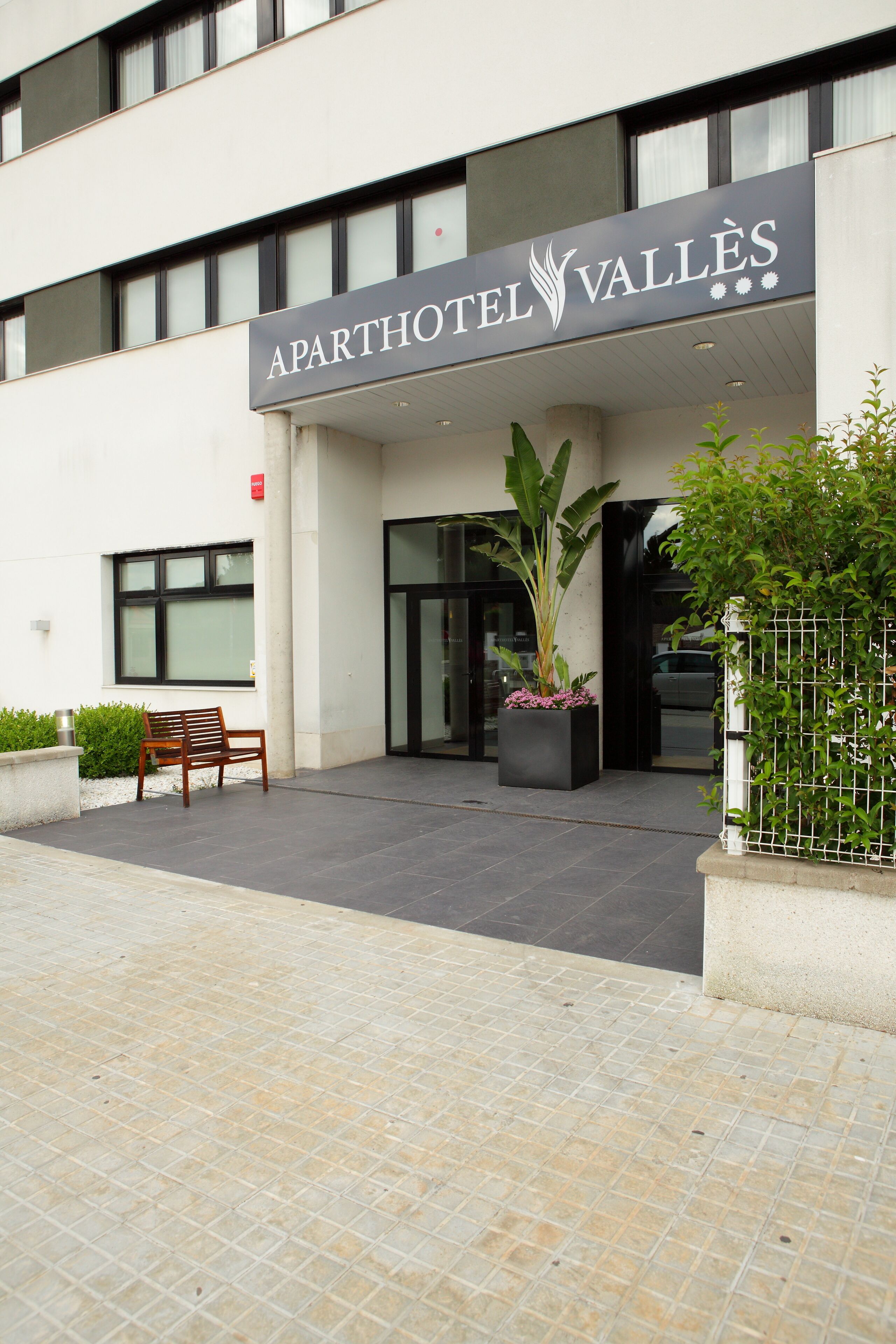 Fasaden på ett lägenhetshotell som heter ”APARTHOTEL VALLÈS”, med en modern arkitektur, en bänk, krukväxter och en grind vid entrén.