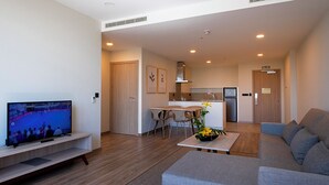 A modern living room with a flat-screen TV, a grey sofa, a coffee table with flowers, a dining area with chairs, and a kitchen with a refrigerator and stove.