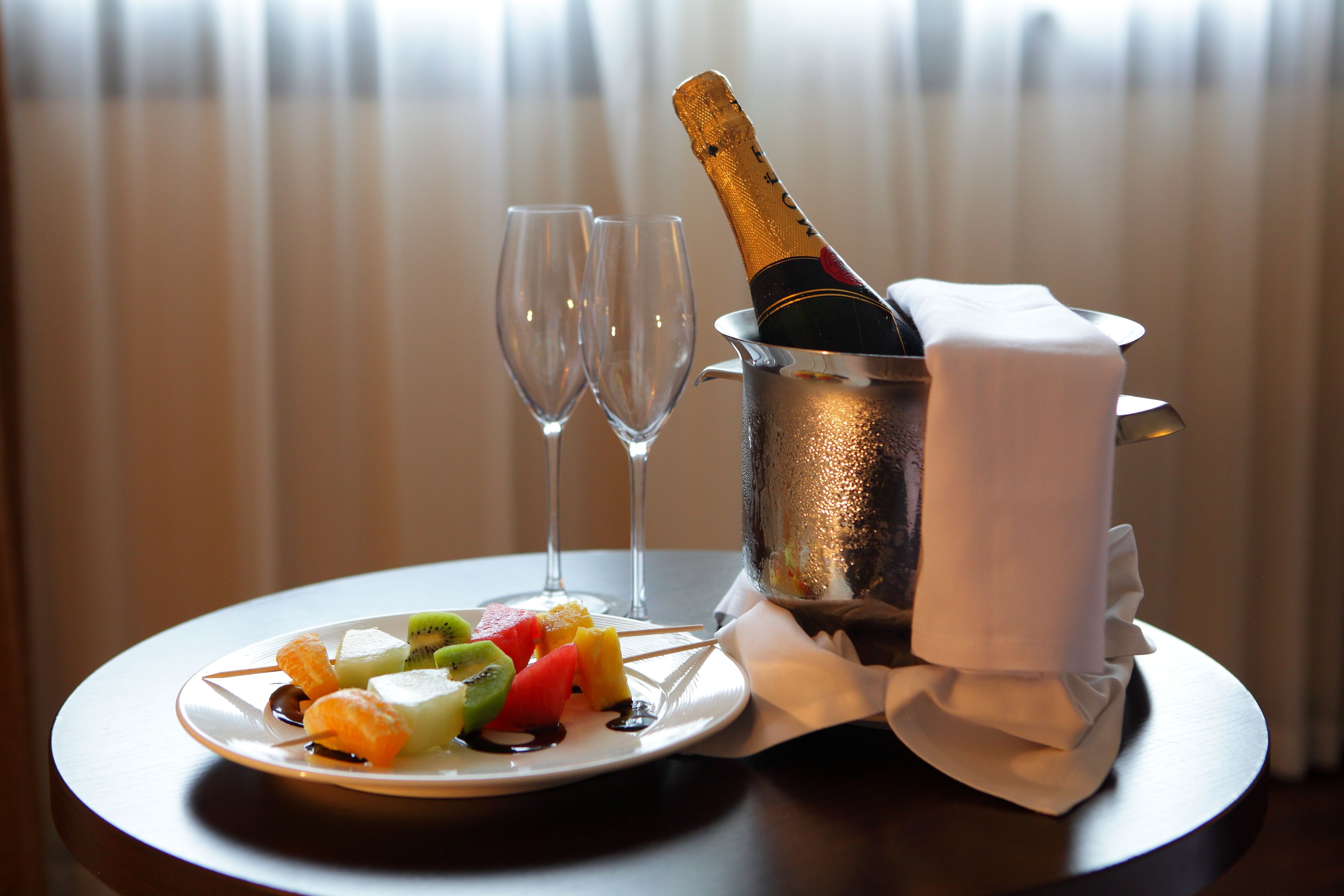 Una mesa con un plato de fruta, dos copas de vino vacías y una botella de champán en un balde de metal.