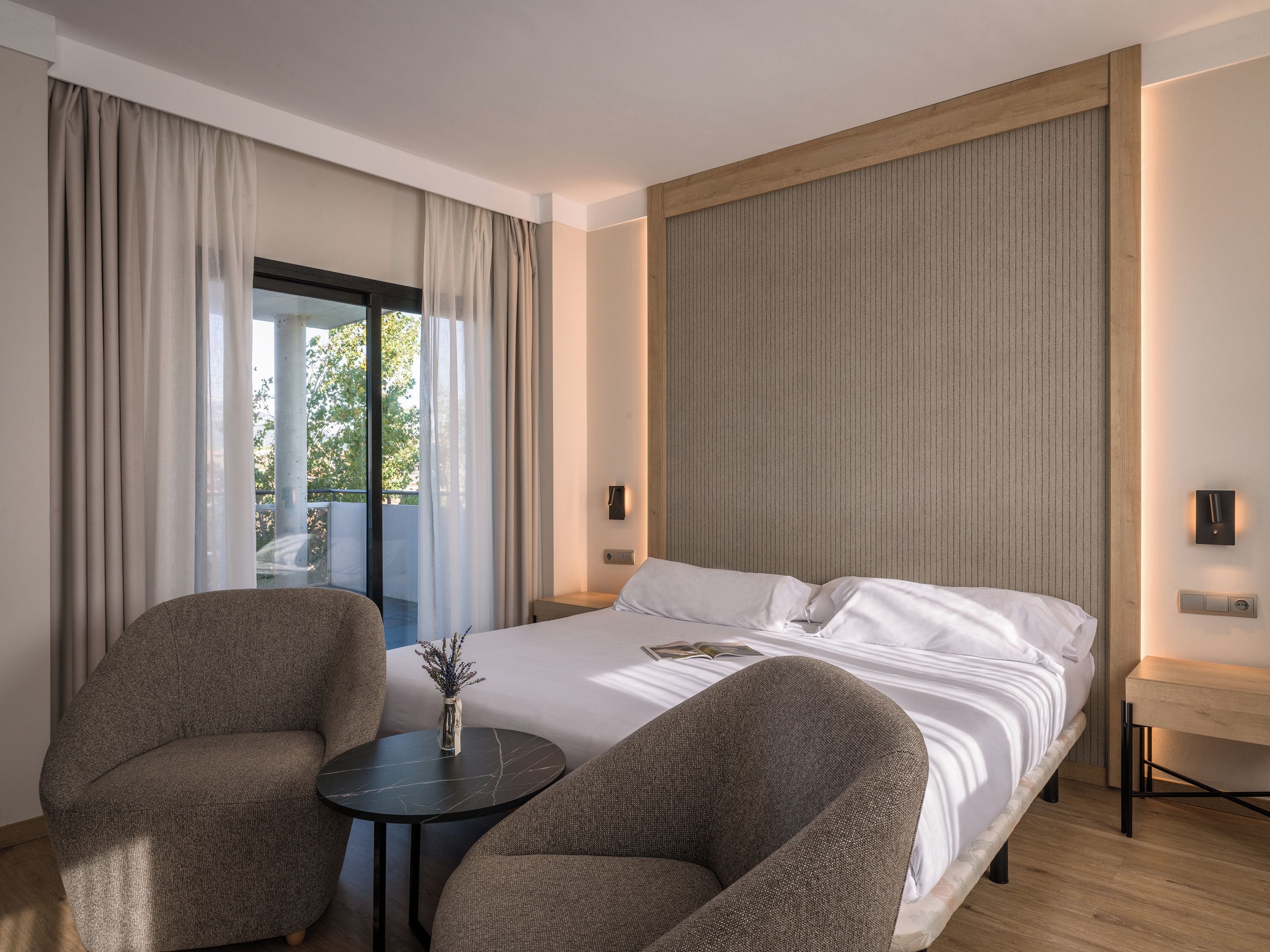 Una habitación de hotel con una cama, dos sillones, una mesita y vistas al exterior.