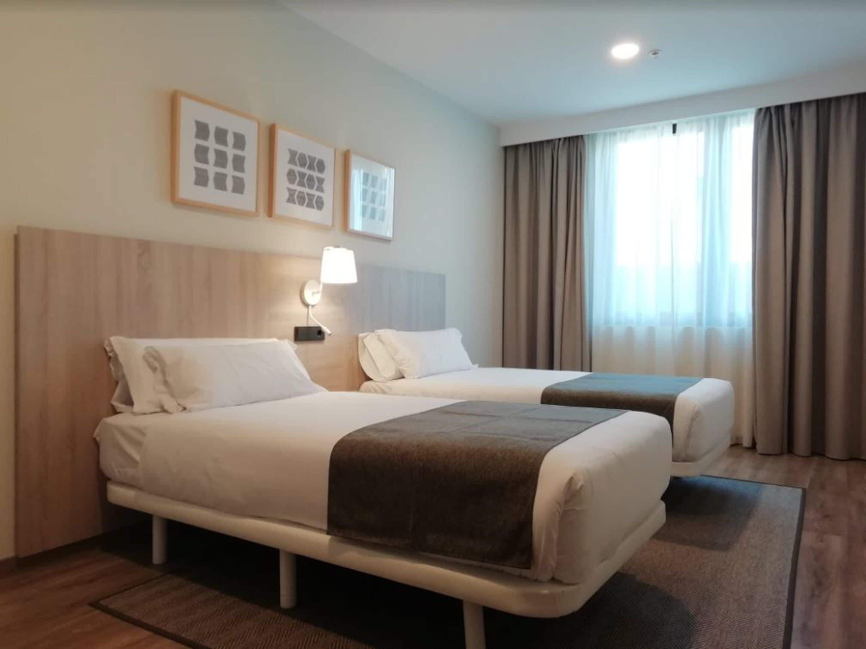 A hotel room with two beds, a bedside table with a lamp, and a window with curtains.