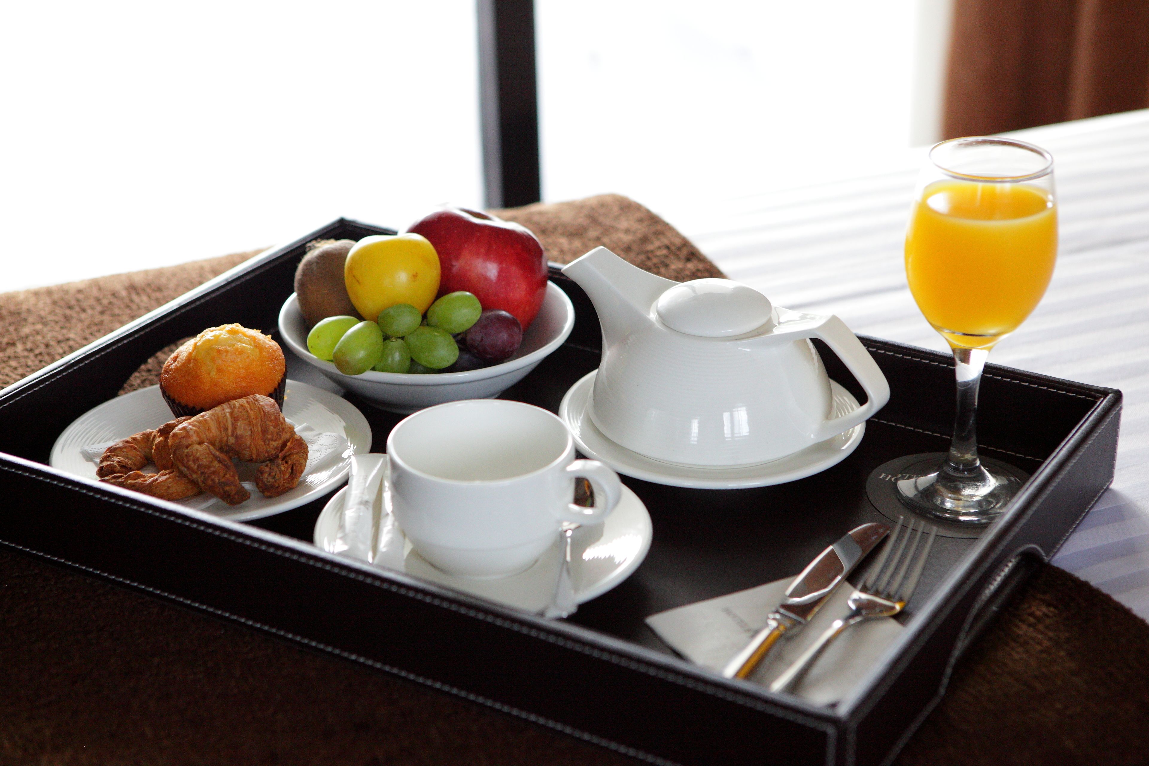 A breakfast tray with croissants, a bowl of fruit, a cup of coffee, a teapot, a glass of orange juice, and cutlery.